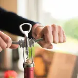 Hände öffnen eine Weinflasche mit einem Korkenzieher. Der korken wird gerade herausgezogen.