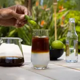 Hand presst Fingerlimette über ein Glas mit Eis, Kaffee und Tonic Water. Ein Krug mit Cold Brew und eine Flasche Tonic stehen daneben.
