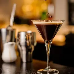 Ein eleganter Cocktail mit Espresso, garniert mit Kaffeebohnen, steht auf einer dunklen Theke. Im Hintergrund sind Barutensilien zu sehen.