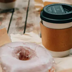 Ein amerikanischer Kaffee im Pappbecher mit braunem Hitzeschutz und ein zuckriger Donut liegen auf einem Holztisch.