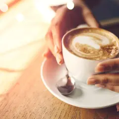 Hände halten eine Tasse cremigen Cappuccinos, der wie ein Kunstwerk mit Latte Art verziert ist. Ein Moment der Ruhe, bevor der erste Schluck den Tag belebt.