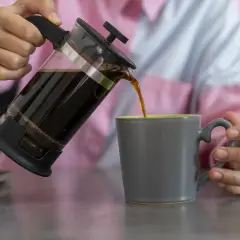 Kaffee wird aus einer French Press in eine graue Tasse gegossen.