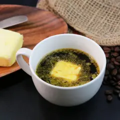 Kaffee mit Butter zum Abnehmen: Ein Block Butter liegt auf einem Holzbrett neben einer Tasse Kaffee mit einer Butterflocke. Geröstete Kaffeebohnen liegen daneben.