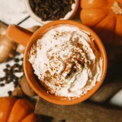 Ein cremiger Pumpkin Spice Latte mit Sahne und Zimt, umgeben von Kürbissen, Ingwer und Gewürzen.