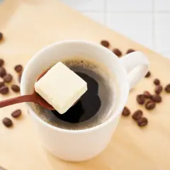 Ein Löffel Butter schwebt über einer Tasse Kaffee, umgeben von Kaffeebohnen auf einem Holztablett. Ein ungewöhnlicher Genuss: Kaffee mit Butter.