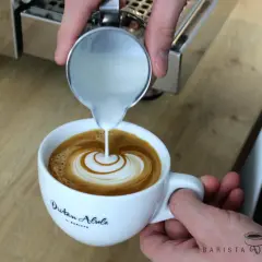 Barista vollendet die Flat White Zubereitung mit kunstvollem Latte Art Gießen. Milch fließt in Espresso und erzeugt ein Herzmuster.