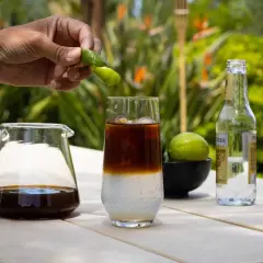 Hand presst Fingerlimette über ein Glas mit Eis, Kaffee und Tonic Water. Ein Krug mit Cold Brew und eine Flasche Tonic stehen daneben.