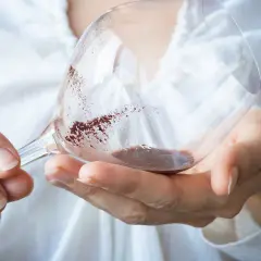 Hände halten ein Weinglas mit Resten von Rotwein. Der Weinstein im Glas schimmert rötlich.