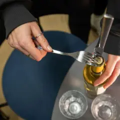Mit einer Gabel wird versucht, eine Weinflasche aufzumachen. Zwei Gläser stehen bereit.