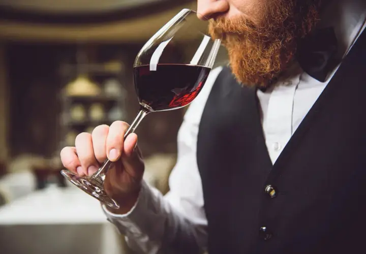 Ein Sommelier mit Bart riecht an einem Glas Rotwein. Er trägt eine Weste und ein Hemd.