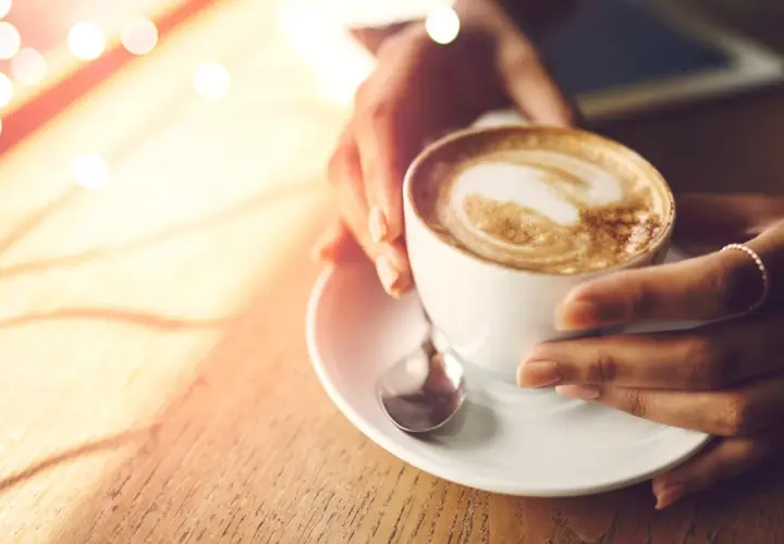 Hände halten eine Tasse cremigen Cappuccinos, der wie ein Kunstwerk mit Latte Art verziert ist. Ein Moment der Ruhe, bevor der erste Schluck den Tag belebt.