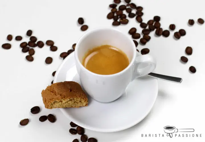Ein cremiger Espresso Lungo in weißer Tasse, daneben ein Cantuccini und Kaffeebohnen.