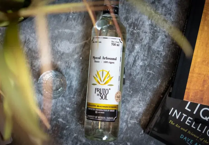 Flasche Mezcal "Fruto del Sol" neben einem Glas und Büchern. So trinkt man Mezcal: pur und langsam genießen.