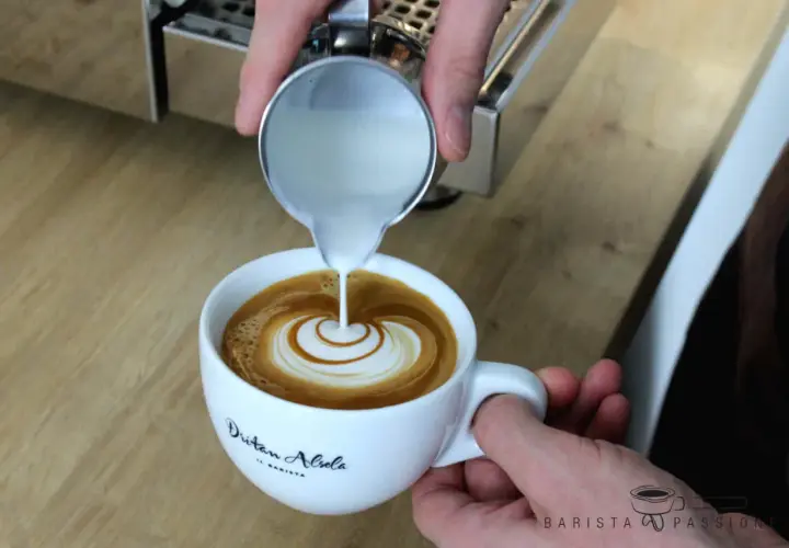 Barista vollendet die Flat White Zubereitung mit kunstvollem Latte Art Gießen. Milch fließt in Espresso und erzeugt ein Herzmuster.
