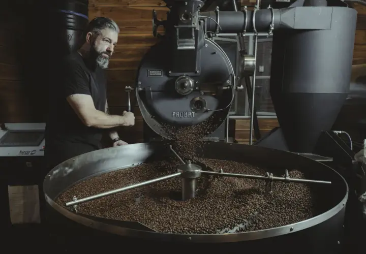 Mann mit Bart bedient Kaffeeröster. Frisch geröstete Bohnen rieseln in die Trommel. Ein Meisterwerk der dritten Welle.