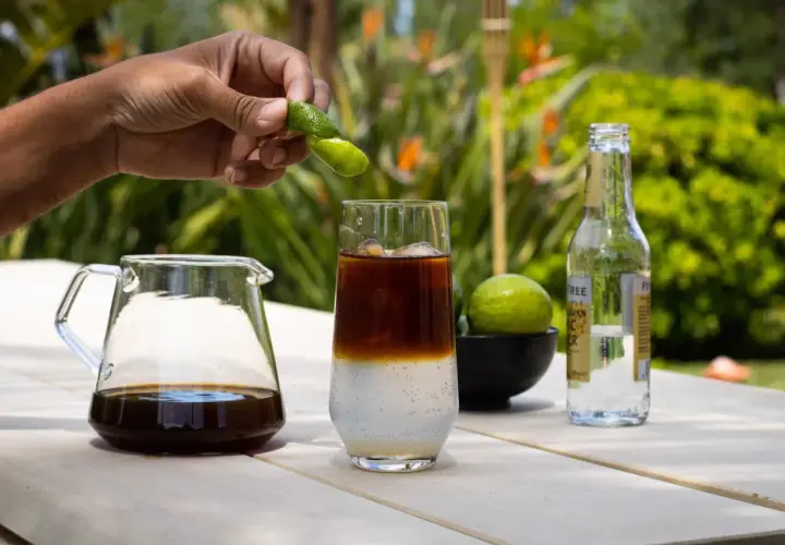 Hand presst Fingerlimette über ein Glas mit Eis, Kaffee und Tonic Water. Ein Krug mit Cold Brew und eine Flasche Tonic stehen daneben.