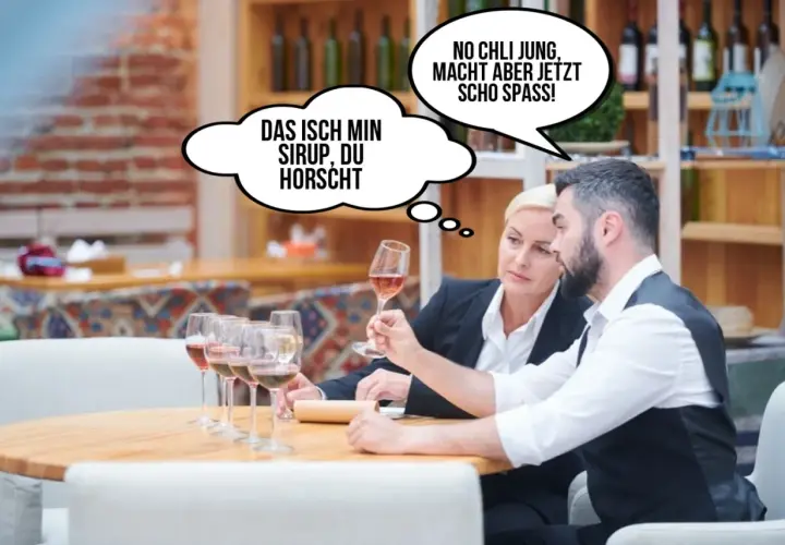 Zwei Personen probieren Wein. Eine denkt: "Das isch min Sirup, du horscht." Die andere: "No chli jung, macht aber jetz scho spass!" Lustig, wie sie den Wein beschreiben.