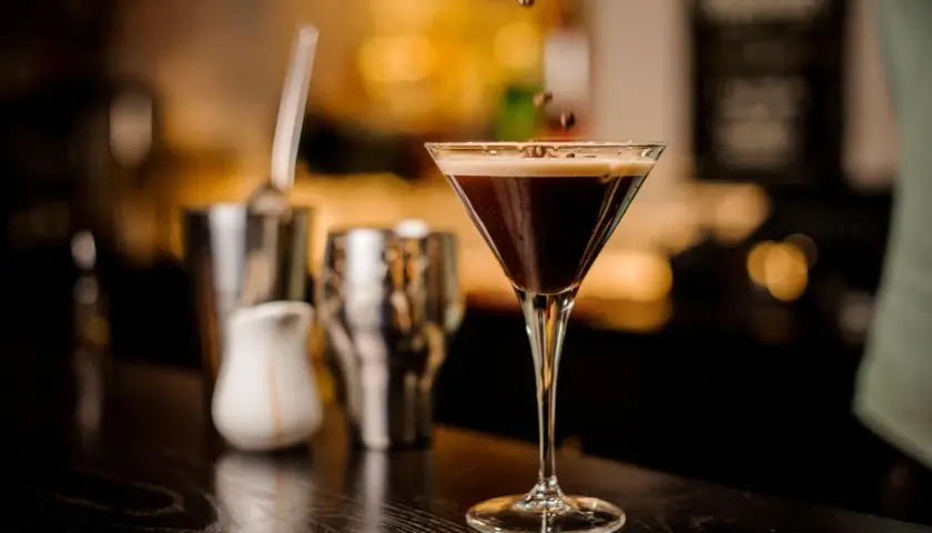 Ein eleganter Cocktail mit Espresso, garniert mit Kaffeebohnen, steht auf einer dunklen Theke. Im Hintergrund sind Barutensilien zu sehen.