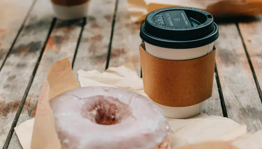 Ein amerikanischer Kaffee im Pappbecher mit braunem Hitzeschutz und ein zuckriger Donut liegen auf einem Holztisch.
