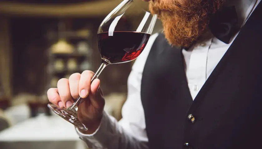 Ein Sommelier mit Bart riecht an einem Glas Rotwein. Er trägt eine Weste und ein Hemd.