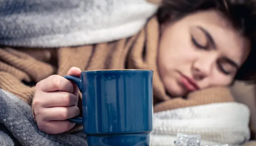 Eine Person mit Halsschmerzen hält eine blaue Tasse Kaffee, eingewickelt in Decken. Medikamente liegen daneben.
