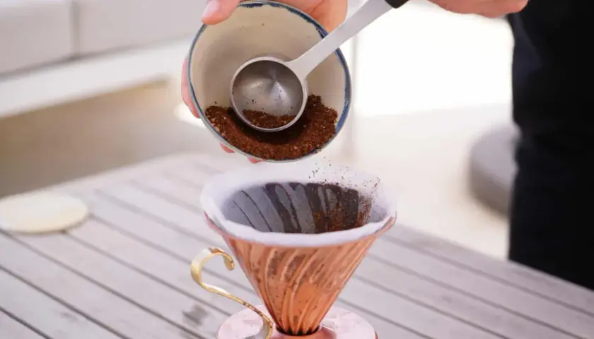 Eine Hand schüttet frisch gemahlenen Kaffee aus einer Schale in einen Kupfer-Filterhalter. So wird der perfekte Kaffee gekocht.