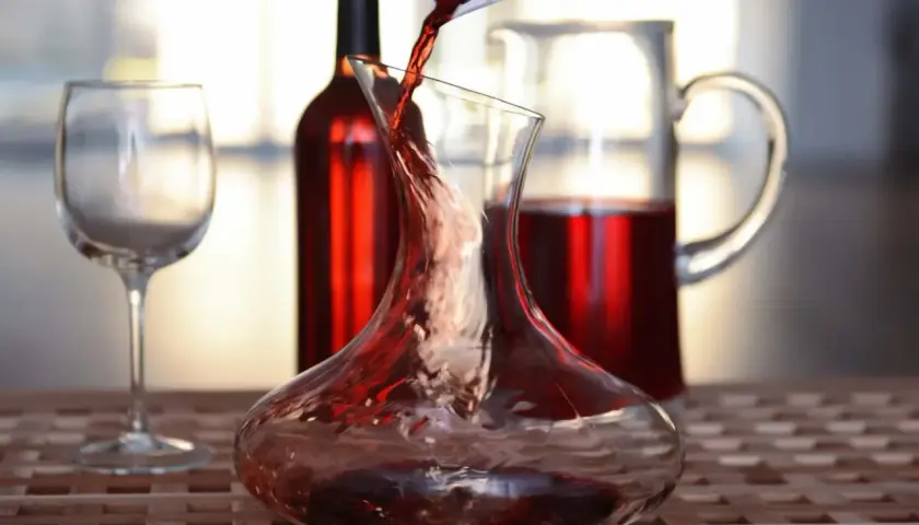 Roter Wein wird in eine Karaffe gegossen, ein Beispiel für Dekantieren. Daneben stehen ein Weinglas und eine weitere Karaffe mit Wein.