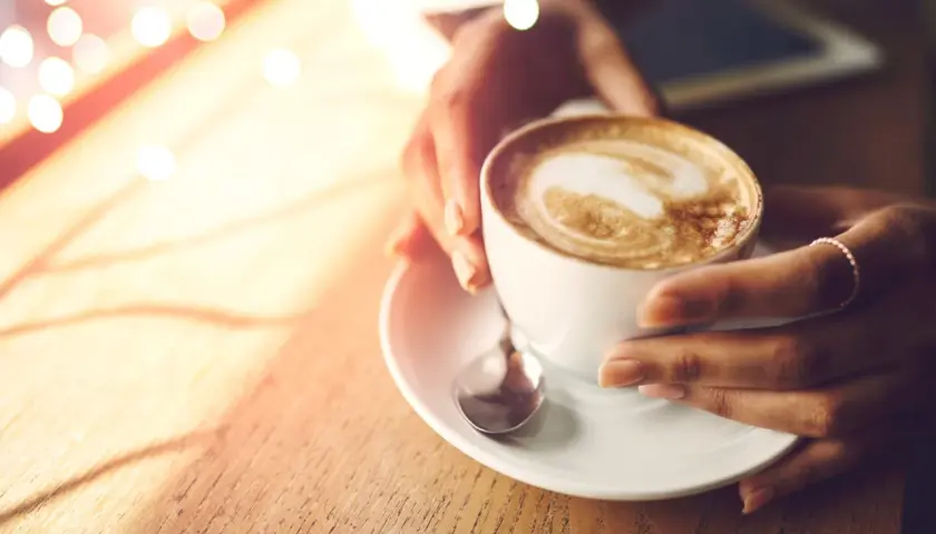 Hände halten eine Tasse cremigen Cappuccinos, der wie ein Kunstwerk mit Latte Art verziert ist. Ein Moment der Ruhe, bevor der erste Schluck den Tag belebt.
