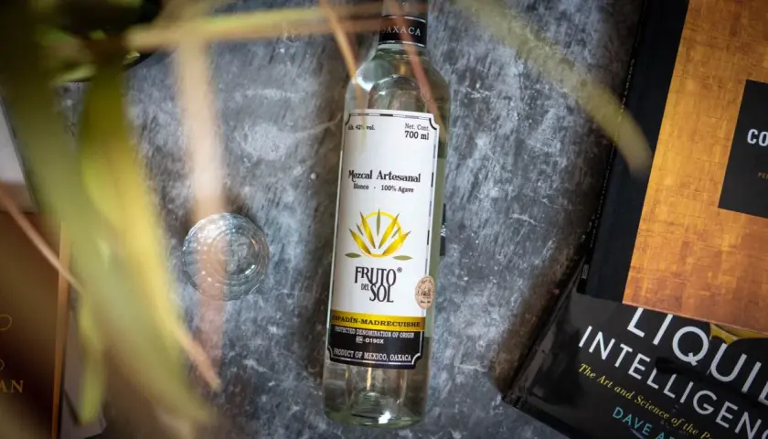 Flasche Mezcal "Fruto del Sol" neben einem Glas und Büchern. So trinkt man Mezcal: pur und langsam genießen.