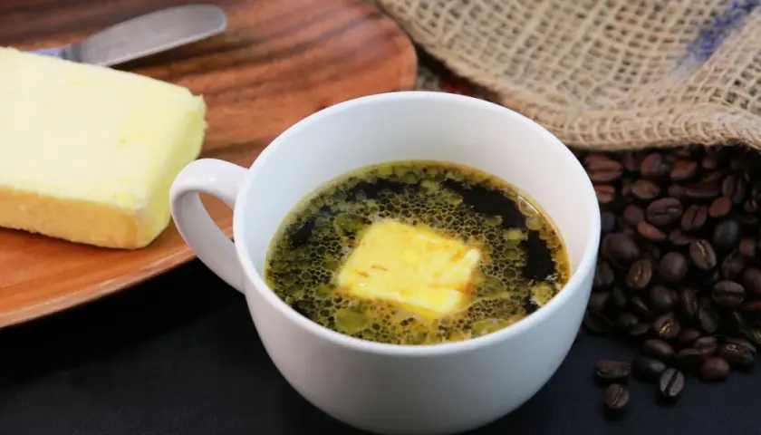 Kaffee mit Butter zum Abnehmen: Ein Block Butter liegt auf einem Holzbrett neben einer Tasse Kaffee mit einer Butterflocke. Geröstete Kaffeebohnen liegen daneben.