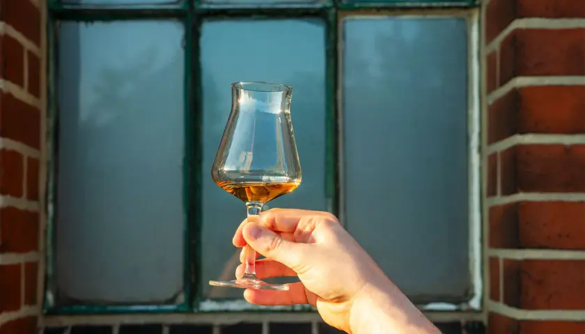 Hand hält ein Glas mit bernsteinfarbenem Whisky. Der Alkoholgehalt des Whiskys wird durch die Farbe und Klarheit angedeutet.
