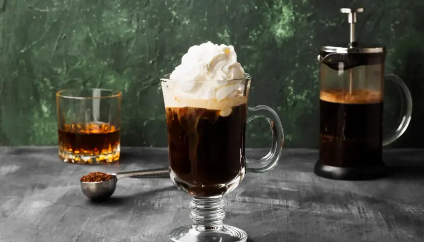 Ein Irish Coffee Rezept: Heißer Kaffee, Whiskey und Sahne in einem Glas, daneben eine French Press und ein Glas Whiskey.