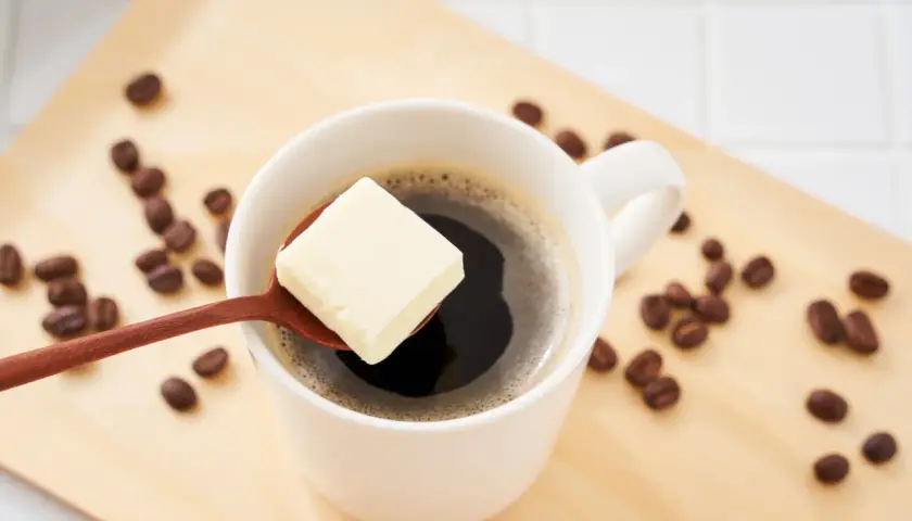 Ein Löffel Butter schwebt über einer Tasse Kaffee, umgeben von Kaffeebohnen auf einem Holztablett. Ein ungewöhnlicher Genuss: Kaffee mit Butter.