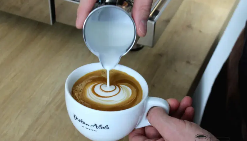 Barista vollendet die Flat White Zubereitung mit kunstvollem Latte Art Gießen. Milch fließt in Espresso und erzeugt ein Herzmuster.