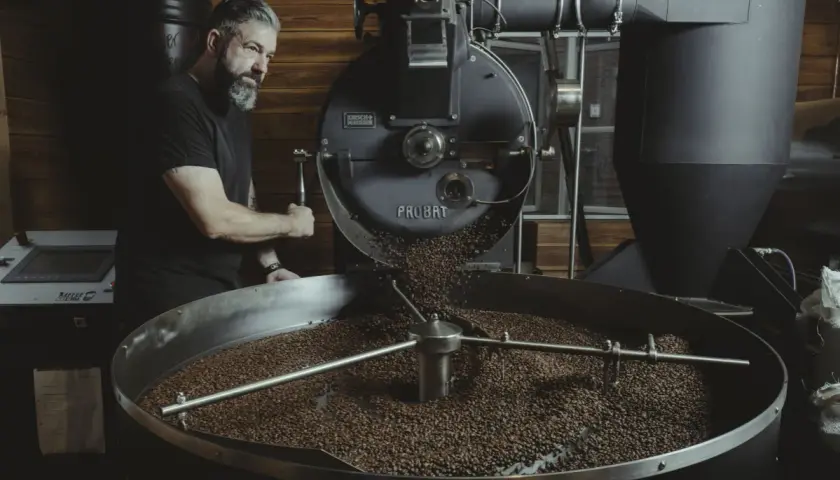Mann mit Bart bedient Kaffeeröster. Frisch geröstete Bohnen rieseln in die Trommel. Ein Meisterwerk der dritten Welle.