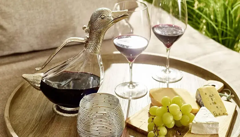 Ein Entenschnabel-Dekanter mit Rotwein, zwei Weingläser, Käse und Trauben auf einem Tablett – so gelingt das Wein richtig einschenken.