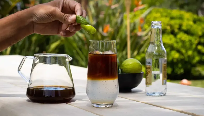 Hand presst Fingerlimette über ein Glas mit Eis, Kaffee und Tonic Water. Ein Krug mit Cold Brew und eine Flasche Tonic stehen daneben.
