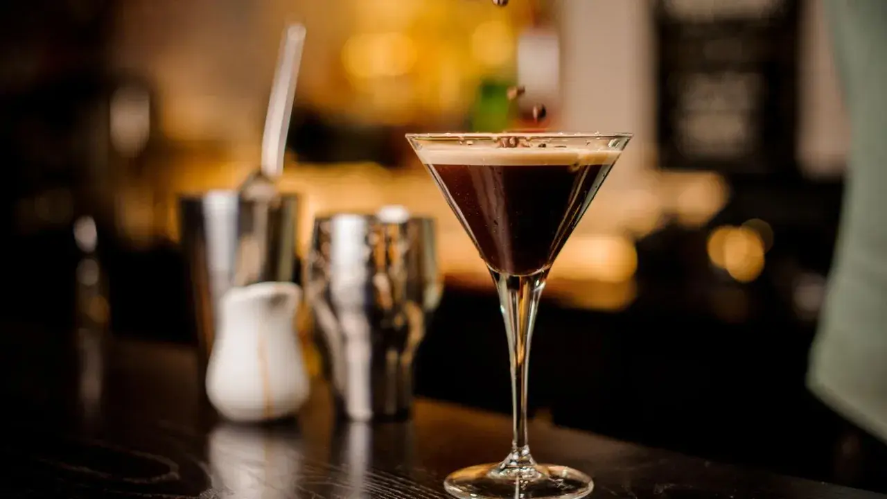 Ein eleganter Cocktail mit Espresso, garniert mit Kaffeebohnen, steht auf einer dunklen Theke. Im Hintergrund sind Barutensilien zu sehen.