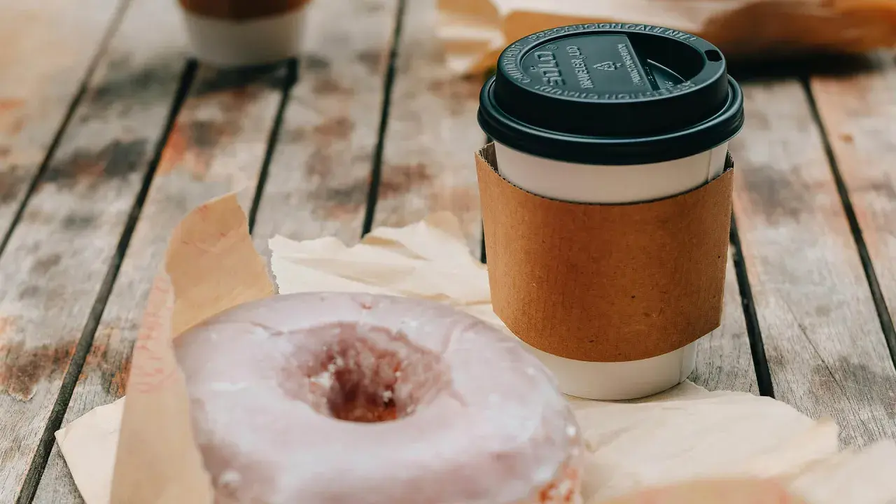 Ein amerikanischer Kaffee im Pappbecher mit braunem Hitzeschutz und ein zuckriger Donut liegen auf einem Holztisch.