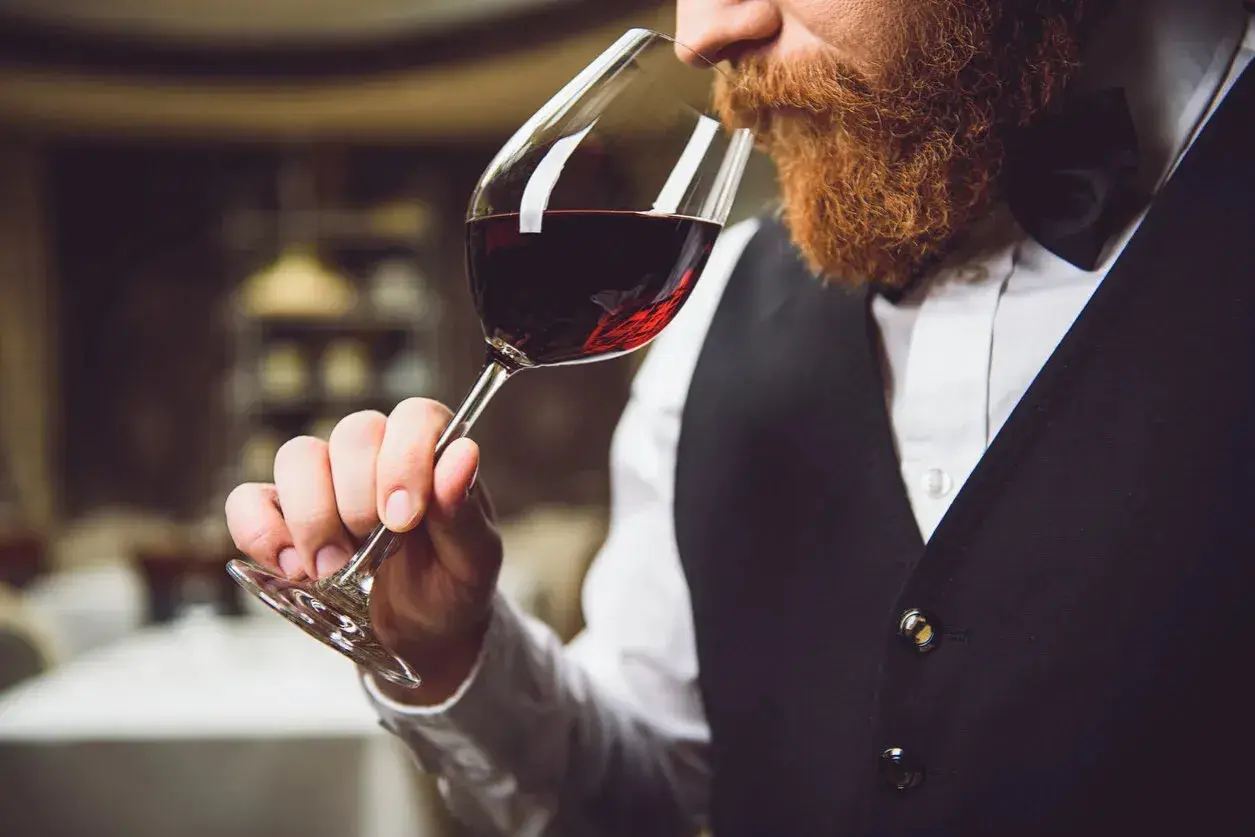 Ein Sommelier mit Bart riecht an einem Glas Rotwein. Er trägt eine Weste und ein Hemd.