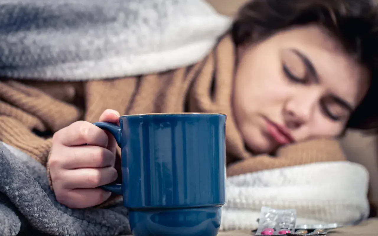 Eine Person mit Halsschmerzen hält eine blaue Tasse Kaffee, eingewickelt in Decken. Medikamente liegen daneben.