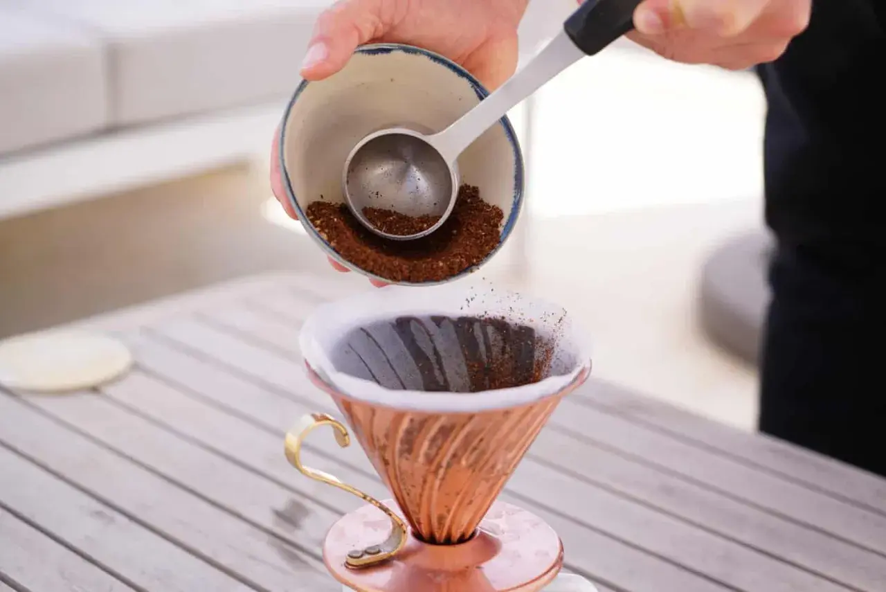 Eine Hand schüttet frisch gemahlenen Kaffee aus einer Schale in einen Kupfer-Filterhalter. So wird der perfekte Kaffee gekocht.