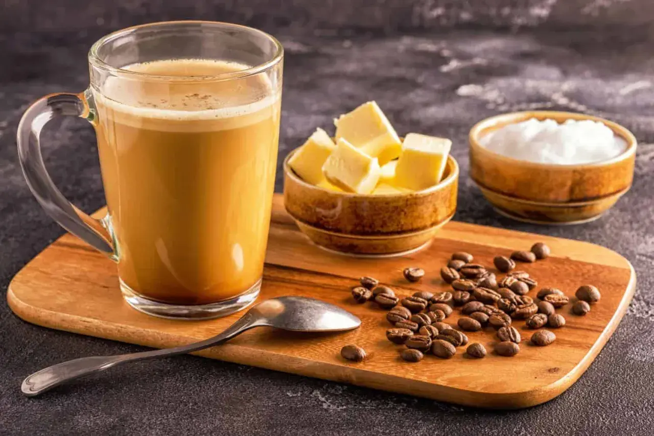 Ein Glas cremiger Kaffee, daneben Butterwürfel und Kokosöl. So wird Bulletproof Coffee zubereitet.
