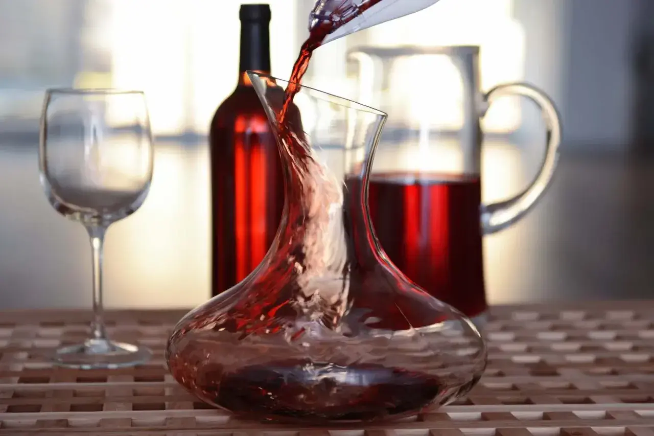 Roter Wein wird in eine Karaffe gegossen, ein Beispiel für Dekantieren. Daneben stehen ein Weinglas und eine weitere Karaffe mit Wein.
