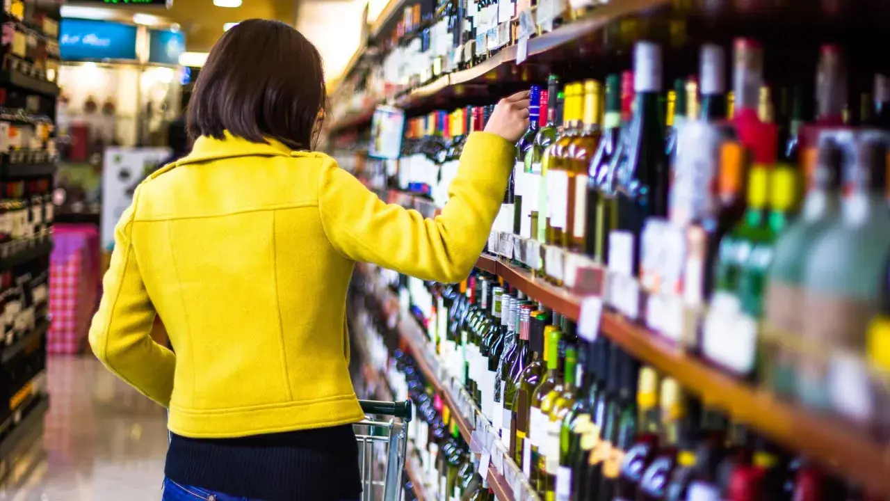 Frau in gelber Jacke wählt Wein aus dem Regal, um die beste **Wein-Bewertung** zu finden.