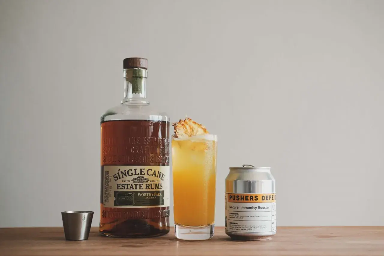 Flasche Single Cane Estate Rums, ein Cocktail mit Ananas und eine Dose "Pushers Defense". Der Rum hat seine Herkunft auf Jamaika.