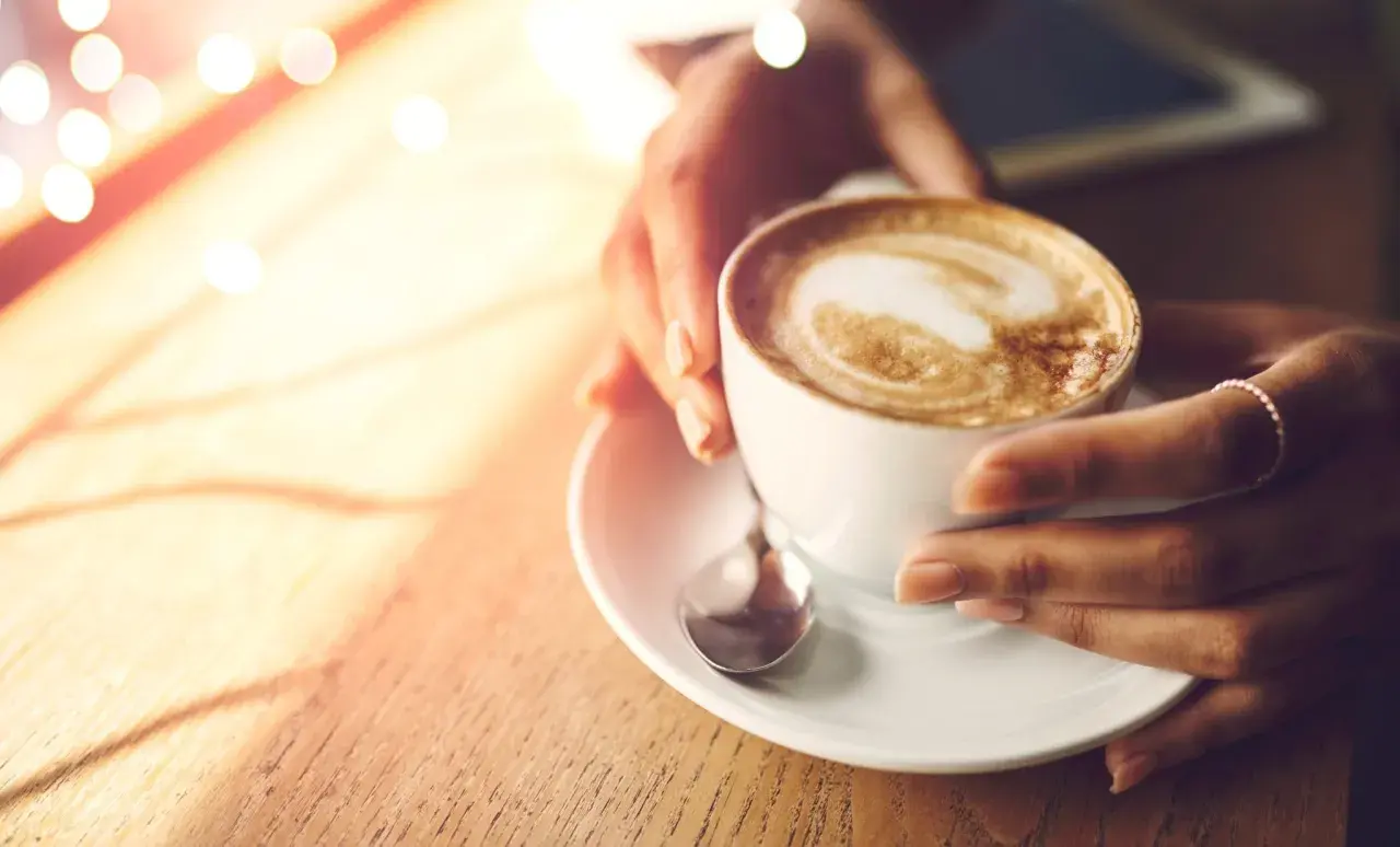 Hände halten eine Tasse cremigen Cappuccinos, der wie ein Kunstwerk mit Latte Art verziert ist. Ein Moment der Ruhe, bevor der erste Schluck den Tag belebt.