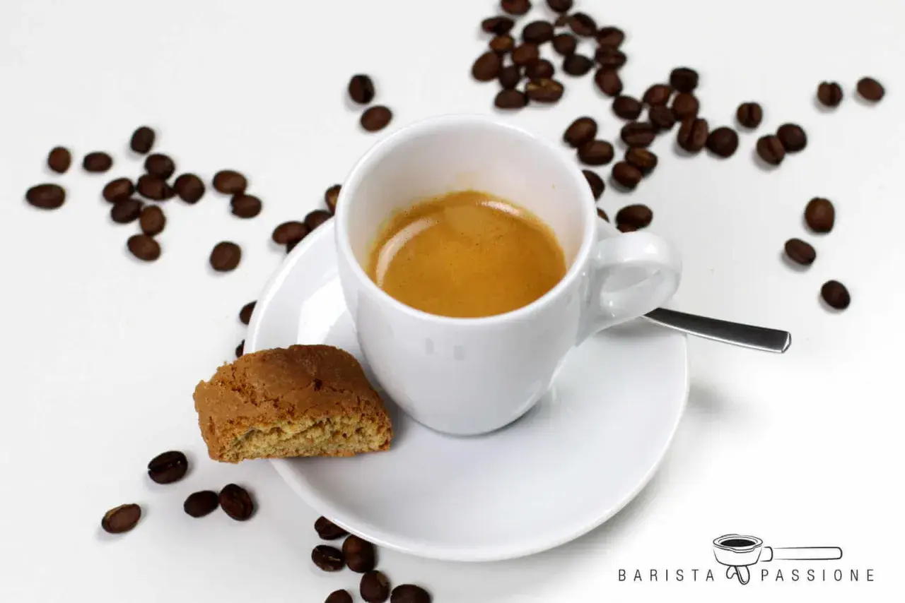 Ein cremiger Espresso Lungo in weißer Tasse, daneben ein Cantuccini und Kaffeebohnen.
