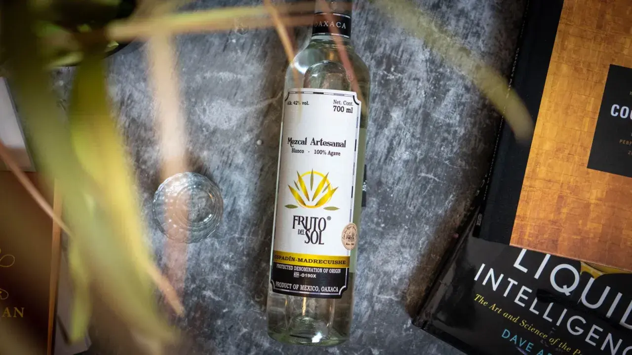 Flasche Mezcal "Fruto del Sol" neben einem Glas und Büchern. So trinkt man Mezcal: pur und langsam genießen.