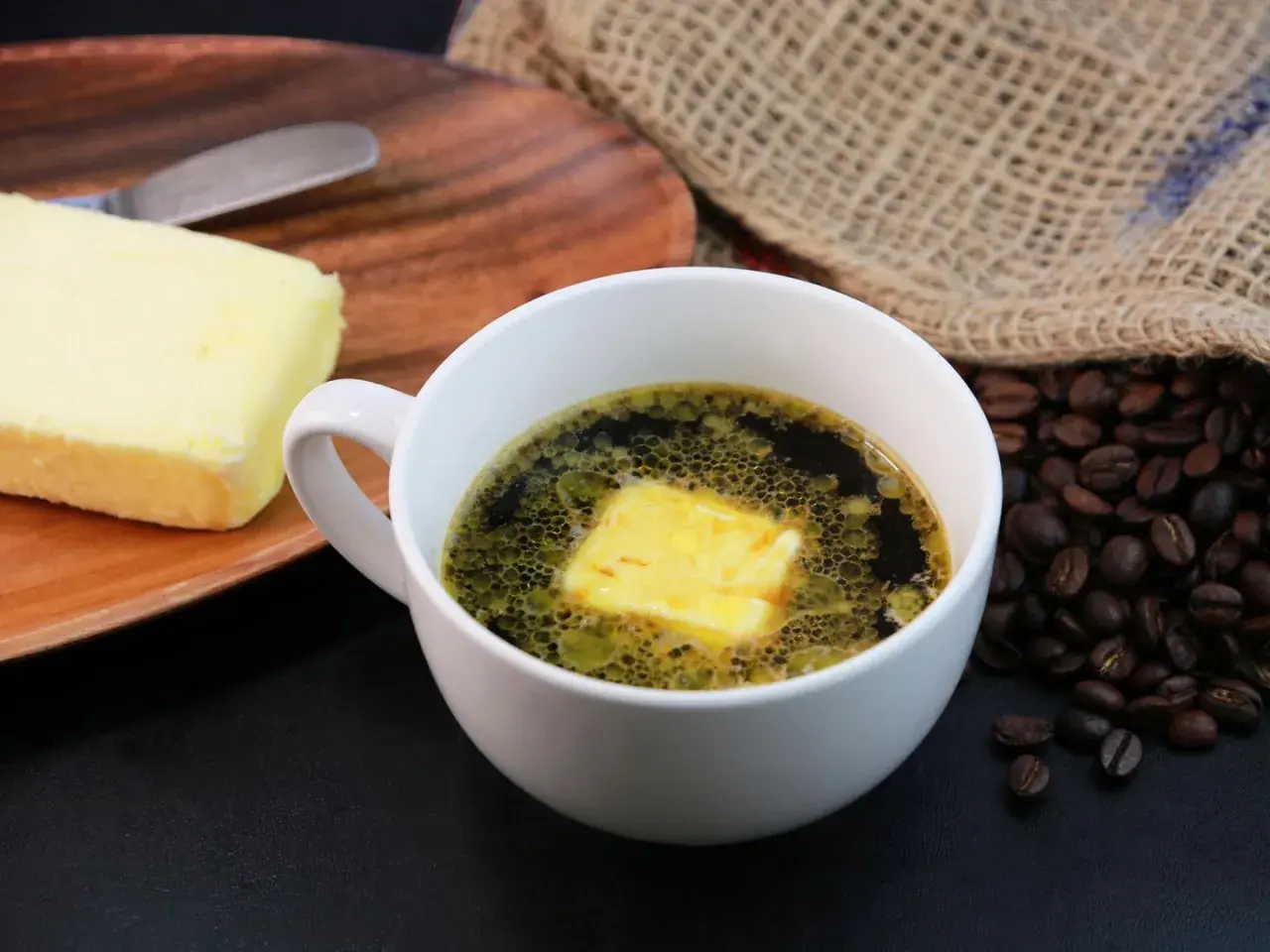 Kaffee mit Butter zum Abnehmen: Ein Block Butter liegt auf einem Holzbrett neben einer Tasse Kaffee mit einer Butterflocke. Geröstete Kaffeebohnen liegen daneben.